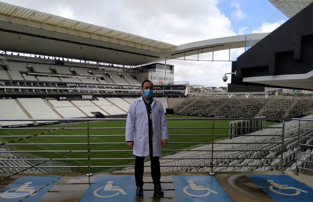 Na imagem está Luiz Artur em pé com jaleco e ao fundo há um estádio de futebol.